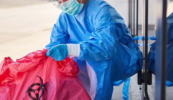 Healthcare worker in protective gown and gloves handling a red biohazard bag