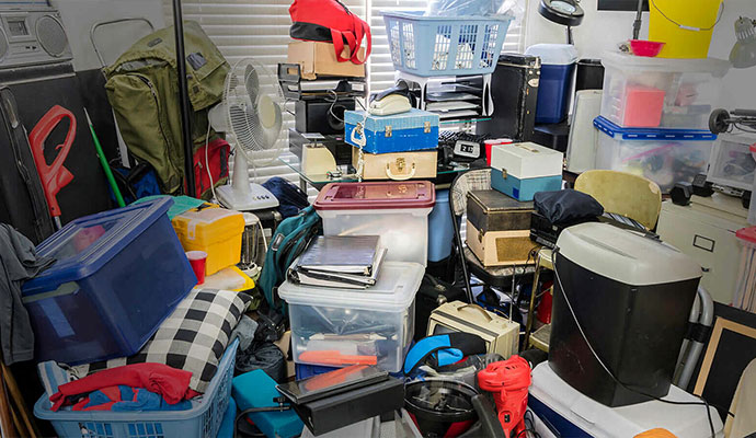 A heavily cluttered room filled with stacked boxes, plastic bins, household items